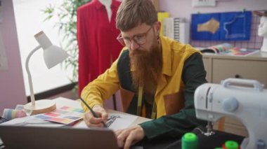 Sakallı bir adam renkli bir terzi stüdyosunda kumaş örnekleri ve dikiş makinesiyle kıyafet tasarımları çiziyor..