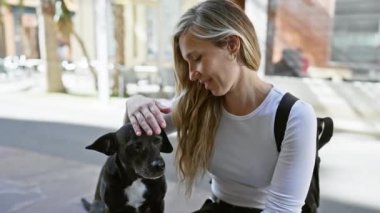 Sevecen genç bir kadın güneşli bir şehir sokağında sakin, siyah labradorunu nazikçe okşuyor, şefkatli bir insan-hayvan bağı sergiliyor.