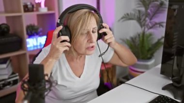 Kulaklık takan odaklanmış bir kadın, iyi ışıklandırılmış bir ev ofisinin oyun bilgisayarında pembe ortam ışıklandırmasıyla oturuyor..