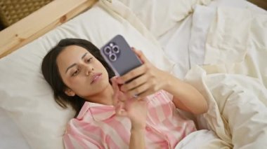 Çizgili pijamalı genç bir İspanyol kadın rahat bir yatak odası ortamında bir yatakta akıllı telefon kullanıyor..