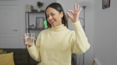 Neşeli, genç, İspanyol bir kadın elinde bir bardak suyla evde gülümseyerek mükemmel bir işaret veriyor.