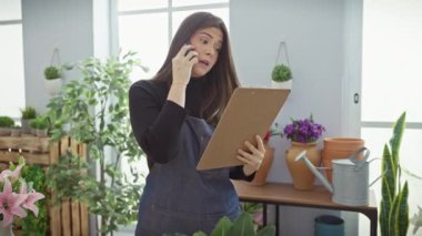 Odaklanmış İspanyol bir kadın, canlı bir çiçekçi dükkanından telefon ve pano ile birden fazla iş yapıyor..