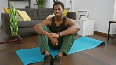 Düşünceli bir adam, modern bir oturma odasındaki yoga minderinin üzerinde, yakınlardaki spor aletleriyle antrenman yapıyor..