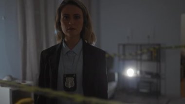 Ciddi bir beyaz kadın dedektif loş ışıkta duran bir suç mahallinde duruyor, polis rozeti görünür..
