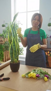 Çiçekçide bitkilerle ilgilenen Afrikalı Amerikalı kadın neşeli bir ifadeyle ve bahçe eldivenleri takıyor..