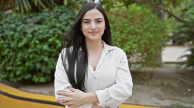 Açık şehir parkında kollarını kavuşturup gülümseyen kendine güvenen İspanyol bir kadının portresi.