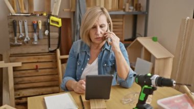 Marangozluk atölyesinde olgun bir kadın marangozluk aletleri arasında dijital tableti izlerken akıllı bir telefonla iletişim kuruyor..