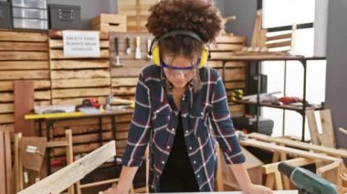 Kıvırcık saçlı ve güvenlik gözlüklü İspanyol kadın marangozluk atölyesinde mutlu bir şekilde çalışıyor..