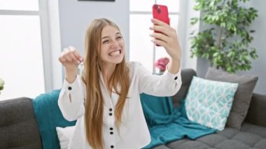 Gıcır gıcır beyaz gömlekli neşeli genç bir kadın yeni bir ev anahtarıyla selfie çekiyor, sıcak bir oturma odasında ev sahipliğini kutluyor..