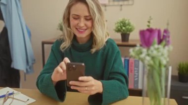 Evde akıllı telefonunu kullanan gülümseyen sarışın bir kadın. Etrafı çiçeklerle ve tahta bir masada gözlüklerle çevrili..