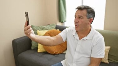Orta yaşlı, beyaz tişörtlü bir adam modern bir oturma odasında video görüşmesi için elinde akıllı telefon tutuyor..
