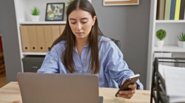 Modern bir ofis içinde dizüstü bilgisayar ve akıllı telefonla çalışan İspanyol bir kadın..