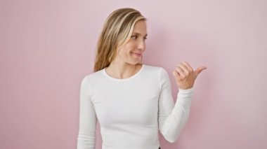 Tişörtlü, pembe izole edilmiş arka planda duran neşeli sarışın kadın. Heyecanlı bir şekilde işaret ediyor ve yan tarafa bakıyor. Başparmak işareti yapıyor. Reklam için mükemmel bir yer.!