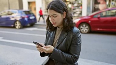 İspanyol kadın şehir caddesinde akıllı telefon kullanıyor. Arka plandaki araç ve dükkanlara odaklanıyor..