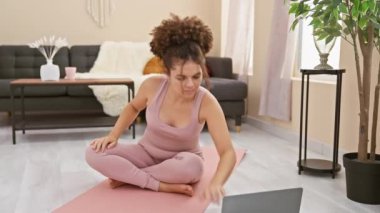 Kıvırcık saçlı, evde yoga yapan genç İspanyol kadın rahat evinde huzurlu bir atmosfer yaratıyor..