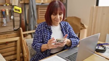Bir ahşap atölyesinde dizüstü bilgisayar ve aletlerle akıllı telefon kullanırken gülümseyen olgun İspanyol kadın..