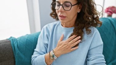 Orta yaşlı, kıvırcık saçlı ve gözlüklü bir kadın oturma odasında kendini rahatsız hissediyor, göğsünü rahatsız tutuyor.