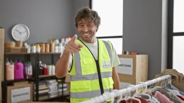 Güvenlik yeleği giymiş neşeli bir erkek gönüllü iyi organize olmuş bir bağış merkezinde kameraya mutlulukla işaret ediyor. Bu da takım çalışması ve şefkat gösteriyor..