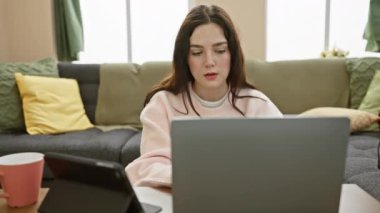 Odaklanmış genç bir kadın evinde laptopuyla rahat bir oturma odasında bir fincan ve tabletle çalışıyor..