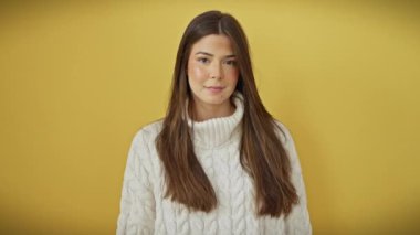 Neşeli genç İspanyol kadın merhaba jesti ile sıcak bir karşılama yapıyor, kazak giyiyor, sarı izole arka planda duruyor, kendine güvenen bir gülümsemeyle el sallıyor.