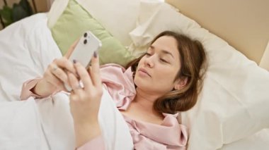 Pembe bluz giyen gülümseyen genç bir kadın, rahat bir yatak odası ortamında, tesadüfen akıllı telefonunu kullanıyor..