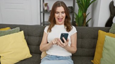Orta yaşlı, kısa saçlı, neşeli bir kadın. Oturma odasında akıllı telefon kullanıyor.