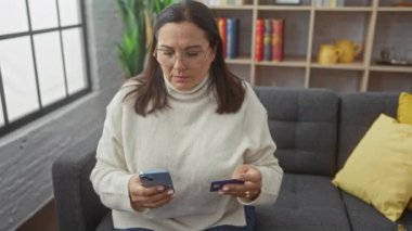 Orta yaşlı İspanyol bir kadın modern bir oturma odasında telefon ve kredi kartına endişeli bir şekilde bakar..