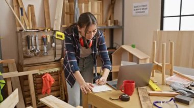 Odun atölyesinde çalışan İspanyol kadın marangoz dizüstü bilgisayarın yanında not defteri yazıyor..
