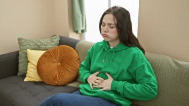 Genç bir kadın, evdeki bir kanepede otururken karın ağrısı çekiyor. Ev ortamında rahatsızlığını dile getiriyor..