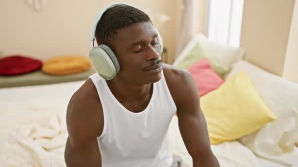 身穿白色油箱上衣的松驰的非洲男人 在五颜六色的舒适卧室里 一边欣赏着耳机 一边欣赏音乐 — 图库视频影像
