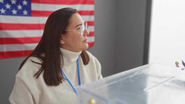 セーターで中年ヒスパニックの女性は 背景に私たちの旗が付いている投票箱を観察します — ストック動画