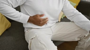 Beyaz elbiseli bir adam gri bir kanepede oturur. Sıcak bir oturma odasında göğsünü acıyla sıkarken.