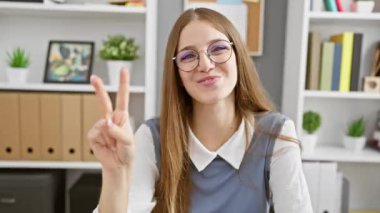 Neşeli genç sarışın iş kadını parmaklarıyla iki numarayı gösteriyor, gülümsüyor ve ofis ortamını işaret ediyor. Güven içinde iletişim kuran neşeli yöneticinin kapalı bir portresi..