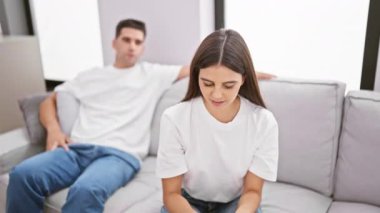 Modern bir oturma odasında, kanepede oturan bir kadın stresli görünürken, arka plandaki adam ilgisiz görünüyor..