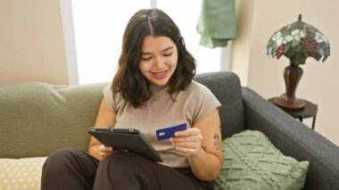 Elinde bir kredi kartı ve tablet olan İspanyol kadın, rahat bir oturma odasında kanepede oturuyor..