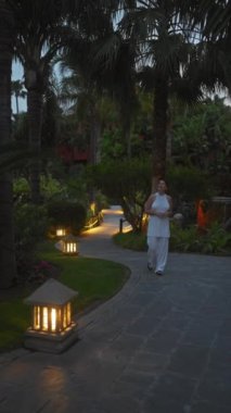 Beyaz elbiseli gülümseyen bir kadın, tropikal palmiyeler ve parlayan yol ışıklarıyla çevrili lüks bir bali otelinde huzurlu bir akşamın tadını çıkarıyor..
