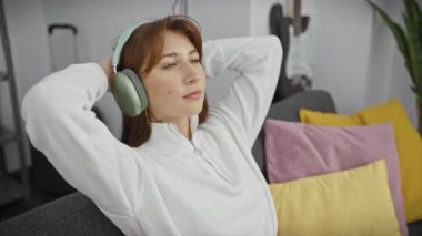 Beyaz kazaklı rahat bir kadın renkli yastıklı bir kanepede yatarken kulaklıkla müzik dinlemekten hoşlanır..