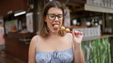 Gözlüklü güzel İspanyol kadın çubukta geleneksel Japon susam topundan zevk alıyor, Kyoto şehrinin otantik sokak yemeği kültürüne daldı.