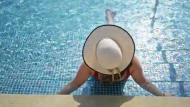 Bir kadın dinlenme yerinin havuzunda geniş kenarlı bir şapkayla uzanır, sakin bir yaz havası uyandırır..