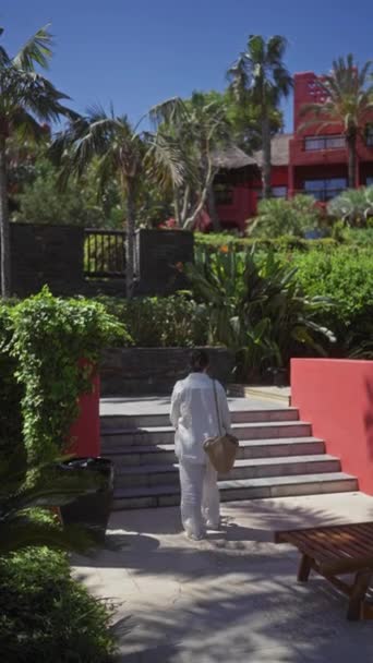 一名身着休闲装的年轻女子在被棕榈树和现代建筑环绕的热带度假胜地走上石阶 — 图库视频影像