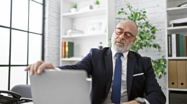 Modern ofis içinde dizüstü bilgisayarla çalışan takım elbiseli olgun sakallı bir adam.