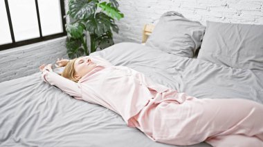 Pembe pijamalı rahat genç bir kadın, parlak yatak odasının iç tarafında gri bir yatak örtüsü olan bir yatağa uzanıyor..