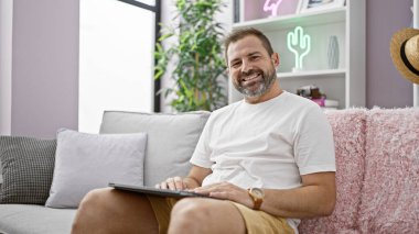 Beyaz saçlı, gülümseyen İspanyol adam koltukta tablet kullanıyor.