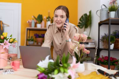 Young caucasian woman florist talking on smartphone using laptop at flower shop