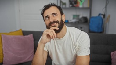 Düşünceli orta yaşlı sakallı bir adam modern bir oturma odasında oturuyor, gülümseyerek yansıtıyor..