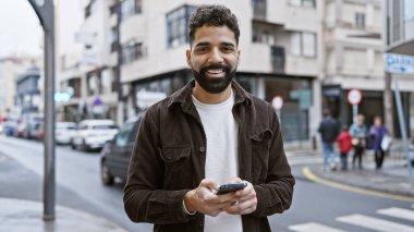 Şehir sokağında arka planda binaları olan akıllı telefon kullanan yakışıklı sakallı adam..