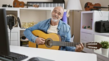 Gülümseyen olgun bir adam müzik aletleri ve kayıt ekipmanlarıyla dolu modern bir ev stüdyosunda gitar çalıyor..