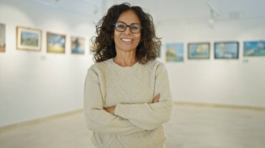Kollarını kavuşturmuş, gülümseyen, olgun İspanyol bir kadın parlak bir sanat galerisinde duruyor..