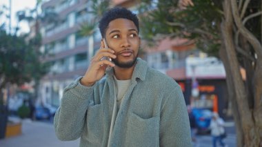 African american man in casual clothing using a smartphone on a busy city street lined with trees and buildings. clipart