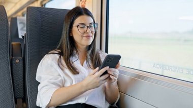 Genç, güzel, İspanyol bir kadın gülümsüyor. Akıllı telefon kullanıyor. Trende oturuyor.